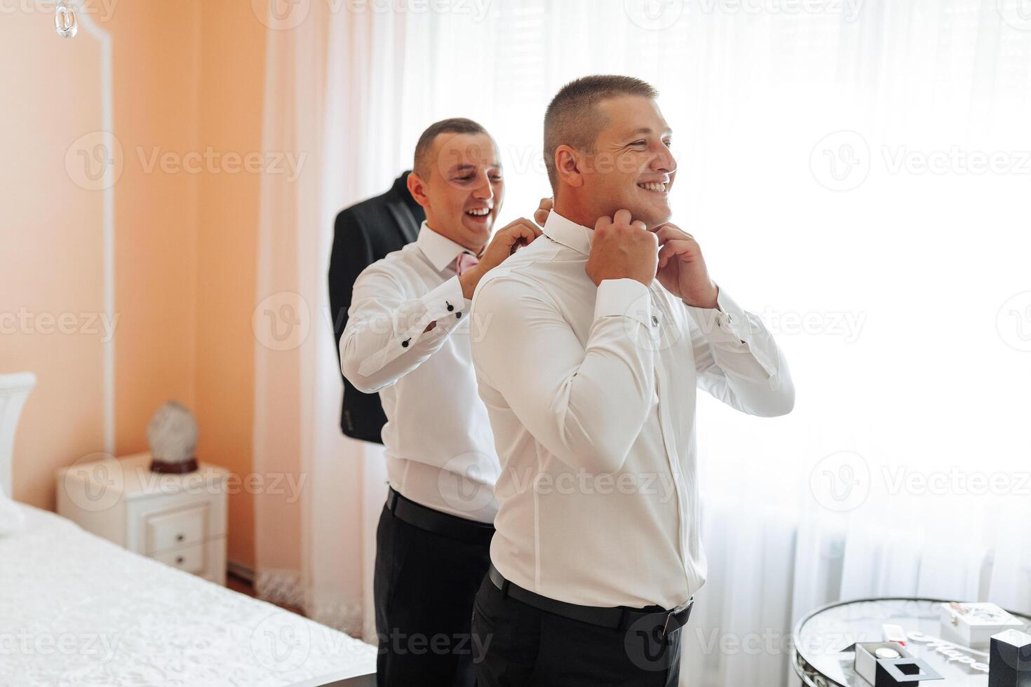 Two men are getting dressed up in a bedroom. One of them is wearing a suit and tie photo