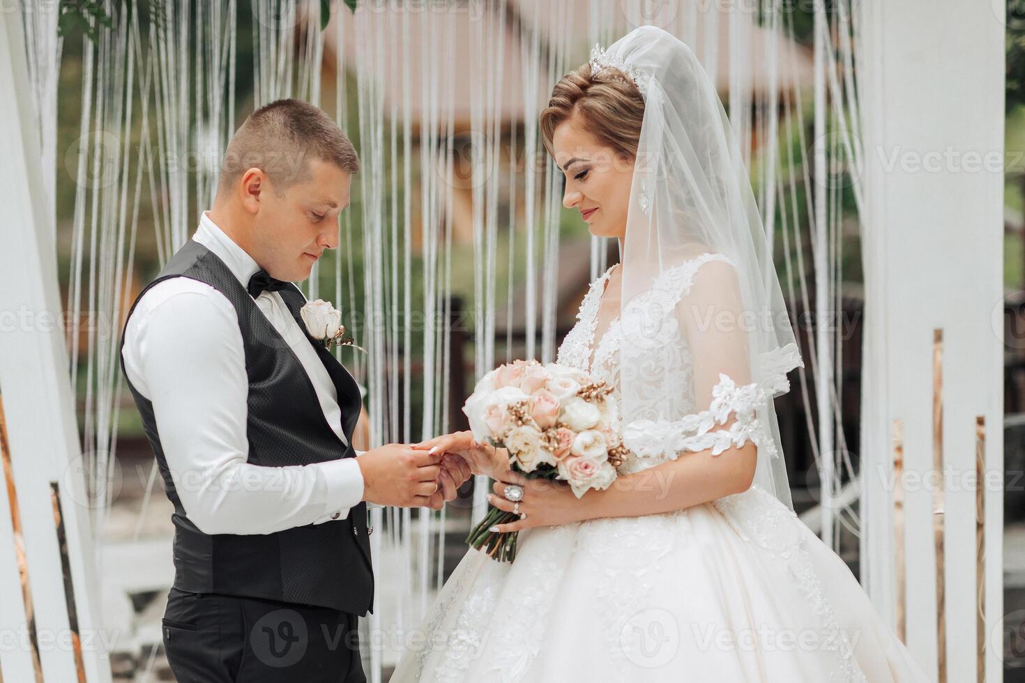 A bride and groom are getting married in a ceremony. The bride is wearing a white dress and the groom is wearing a black suit. They are standing in front of a white curtain photo