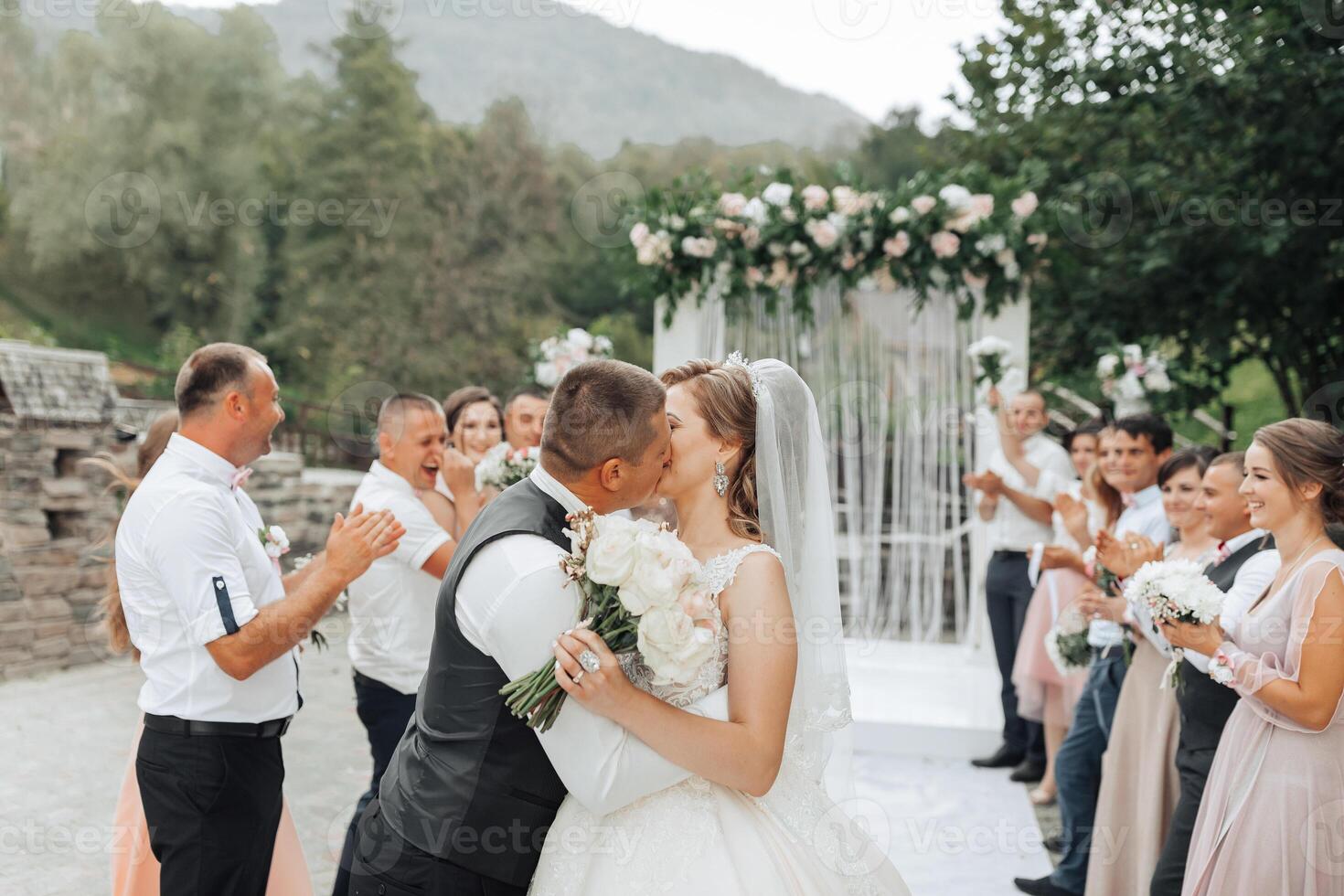 A bride and groom are kissing in front of a crowd of people. The bride is wearing a white dress and the groom is wearing a black vest. The wedding guests are clapping and cheering for the newlyweds photo