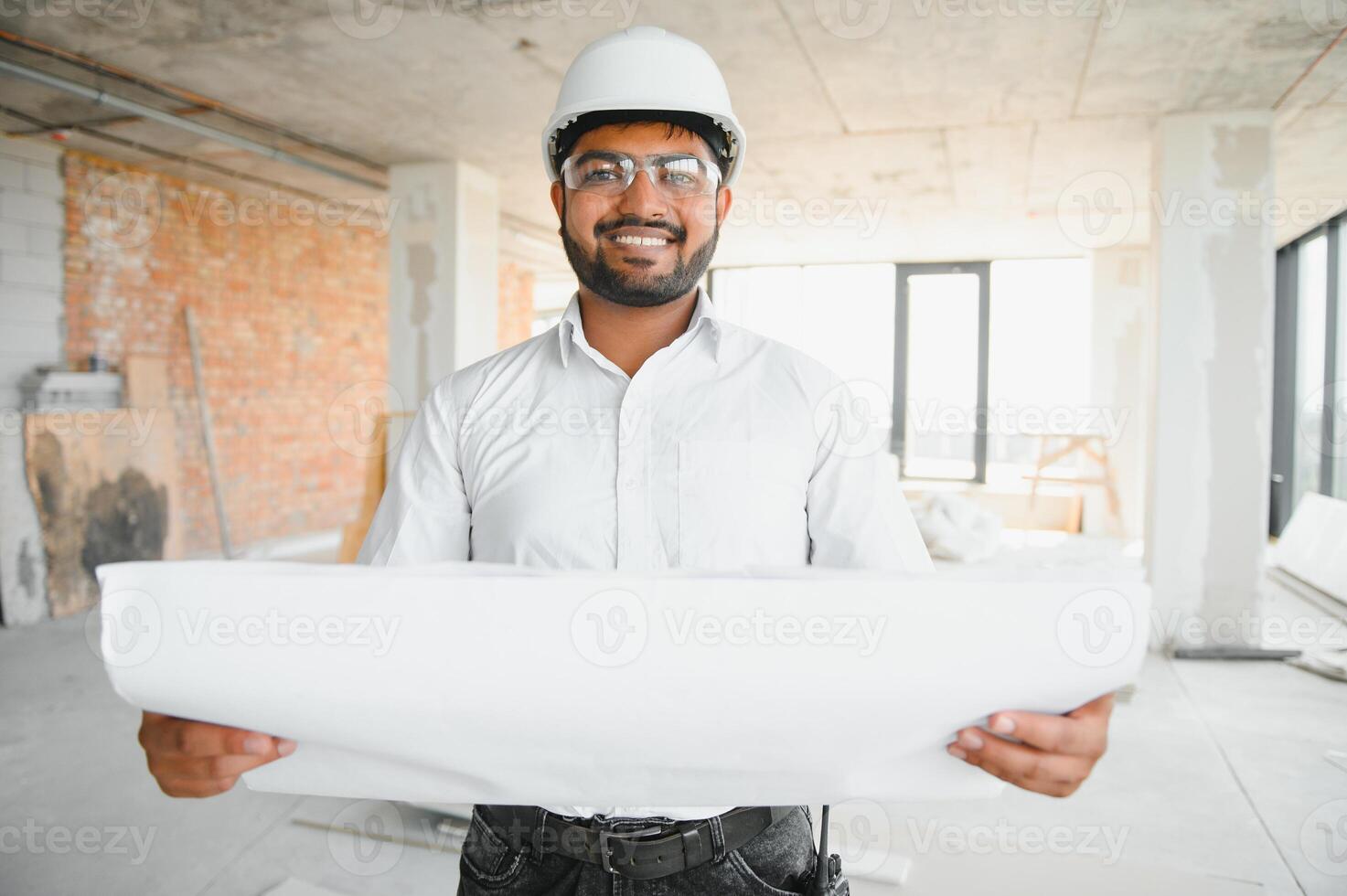 Portrait of young handsome Indian male civil engineer or architect wearing helmet at construction site photo
