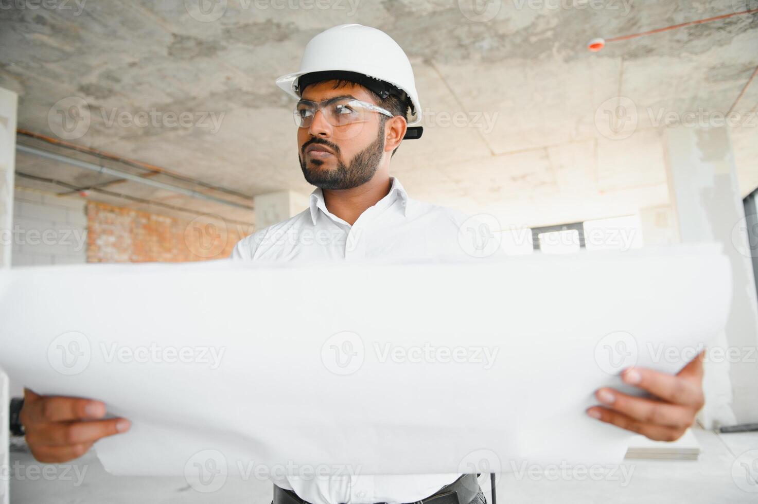 Indian construction site manager standing wearing helmet, thinking at construction site. Portrait of mixed race manual worker or architect photo