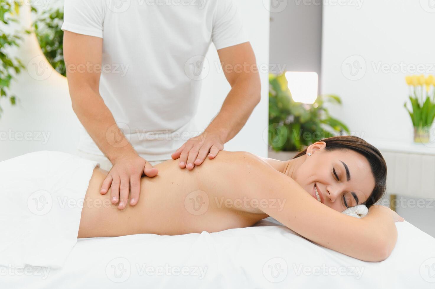Woman enjoying professional body massage at a modern spa salon. Relaxed young lady lying on a massage table while a skilled masseur is gently massaging her back. Beauty, health, pleasure, concept photo