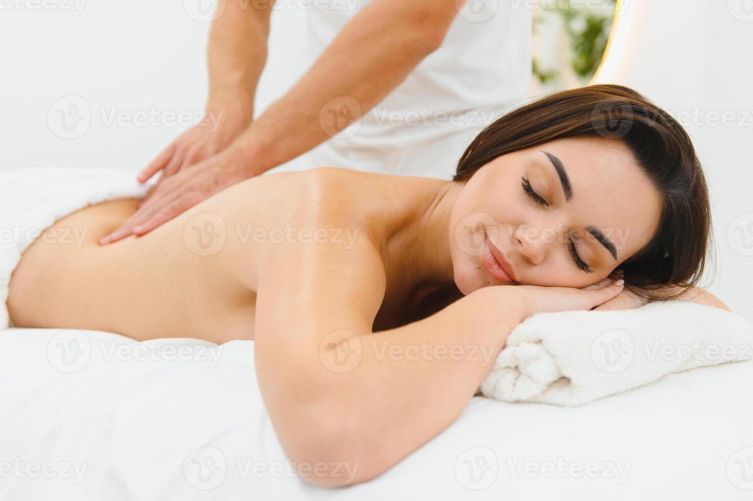Woman enjoying professional body massage at a modern spa salon. Relaxed young lady lying on a massage table while a skilled masseur is gently massaging her back. Beauty, health, pleasure, concept photo