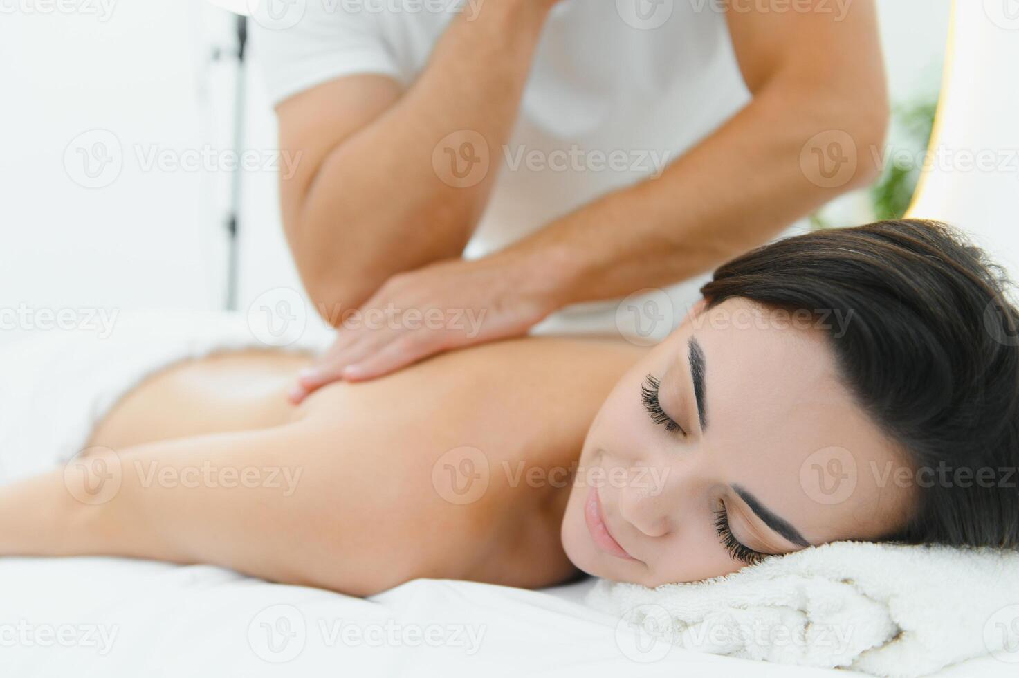 Woman enjoying professional body massage at a modern spa salon. Relaxed young lady lying on a massage table while a skilled masseur is gently massaging her back. Beauty, health, pleasure, concept photo