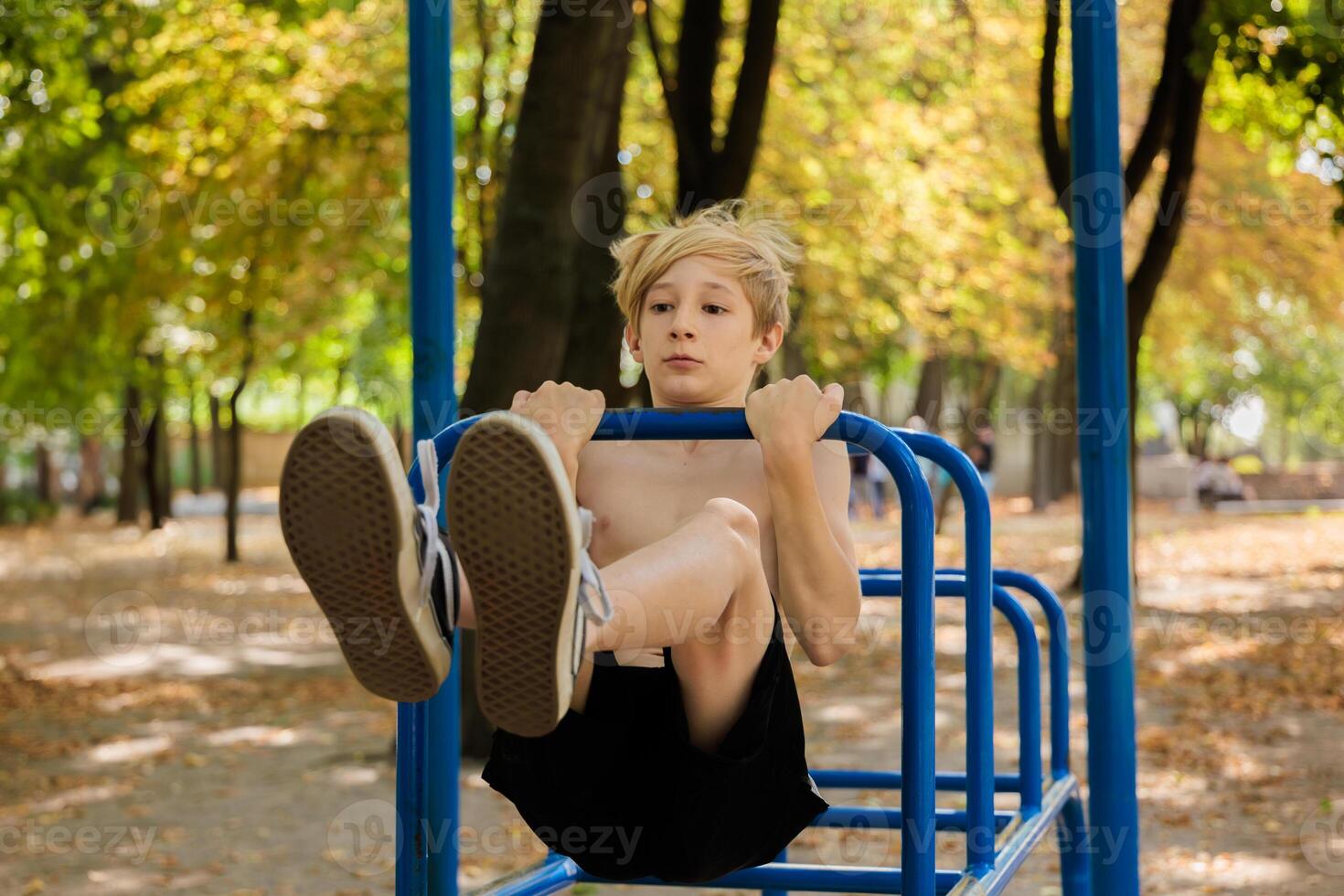 The boy with effort performs a pull-up on the bar with his legs raised in front of him. Athletic teenage boy in the park outdoor workout session photo