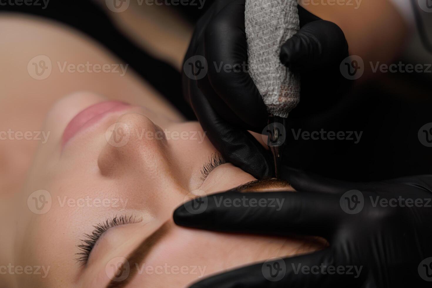 Relaxed girl with closed eyes lying down for permanent makeup procedure as master applies tattoo ideas on eyebrows using tools. Permanent eyebrow makeup, eyebrow tattooing with brown pigment for PMU. photo
