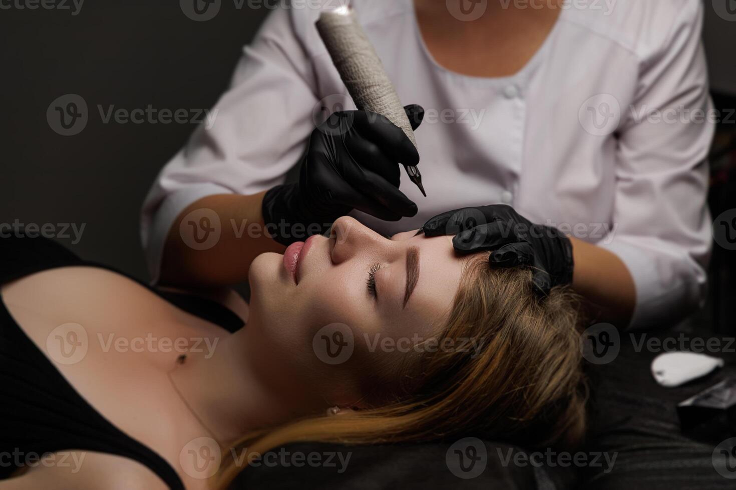 The technician applies precise strokes of pigment to enhance the client's eyebrows, showcasing a delicate beauty procedure in progress. Permanent eyebrow makeup, eyebrow tattooing with brown photo