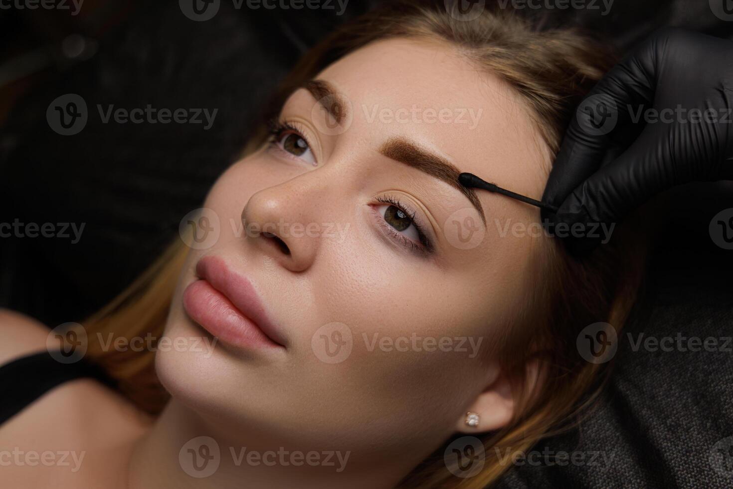 Performed work on permanent makeup for a young girl who lies with open eyes cosmetology salon. Permanent eyebrow makeup, eyebrow tattooing with brown pigment for PMU. photo