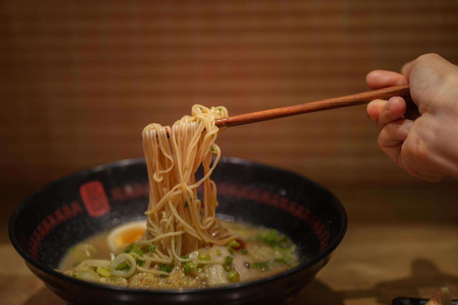 Crop anonymous female with chopsticks eating yummy ramen soup with noodles and meat in black bowl at ramen restaurant. photo