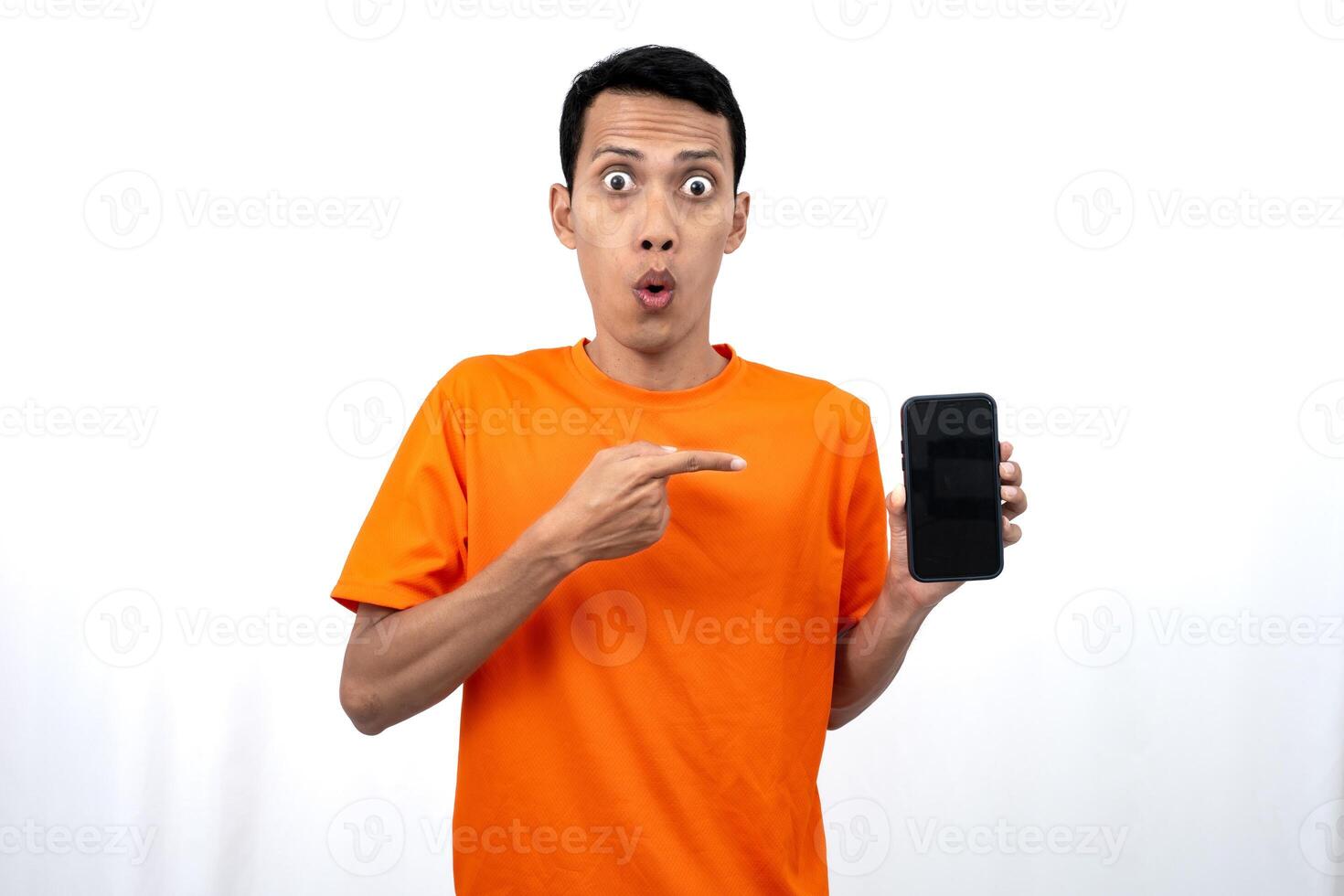 Portrait of a young Asian man wearing an orange t-shirt with a happy, surprised, pleased expression while pointing at a smartphone screen. Isolated on a white background. photo