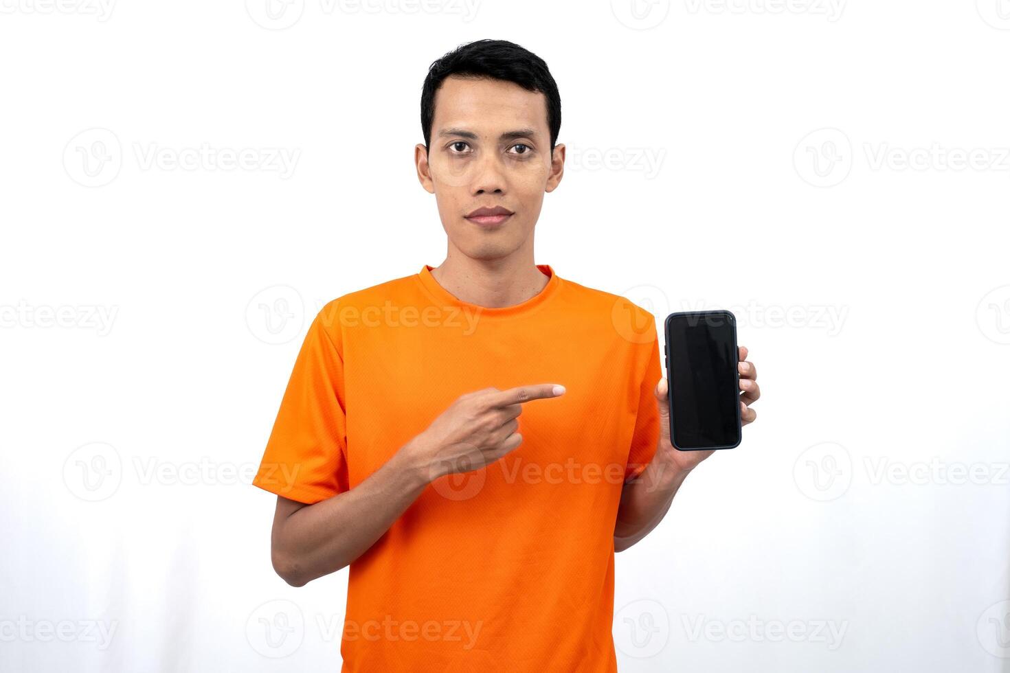 Portrait of a young Asian man wearing an orange t-shirt with a happy, surprised, pleased expression while pointing at a smartphone screen. Isolated on a white background. photo
