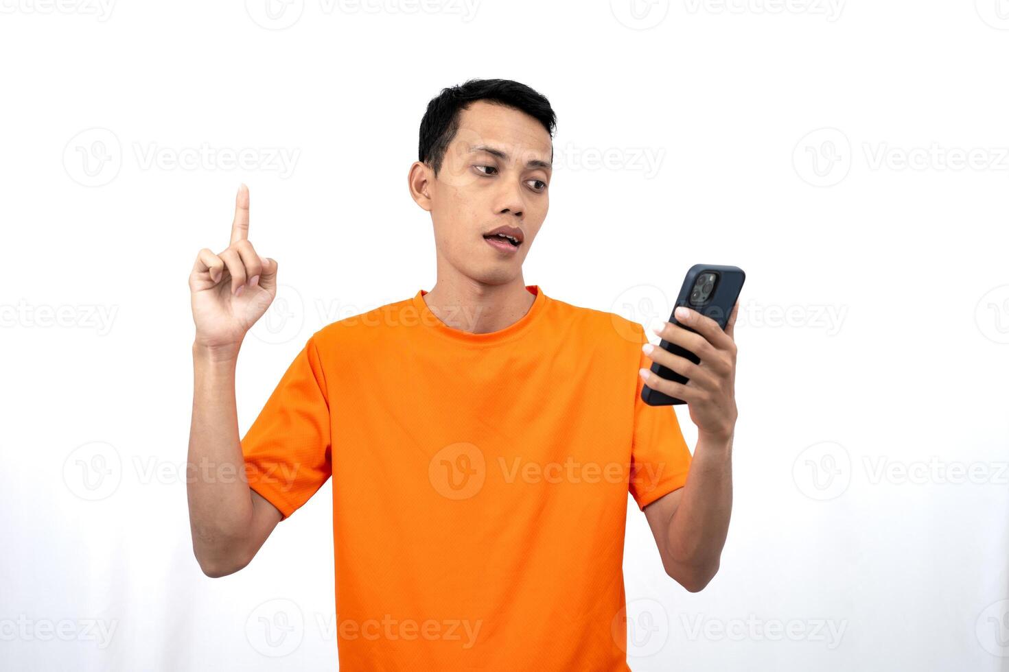 Portrait of a young Asian man wearing an orange t-shirt showing thinking, while holding a smartphone. Isolated on white background. photo