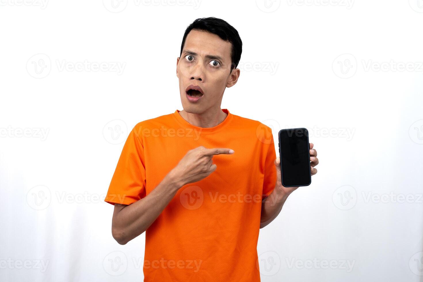 Portrait of a young Asian man wearing an orange t-shirt with a happy, surprised, pleased expression while pointing at a smartphone screen. Isolated on a white background. photo