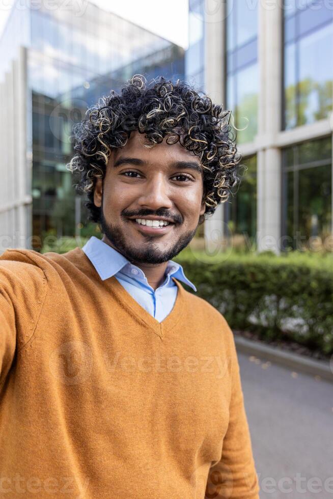 Smiling man taking selfie outside modern office building. Displays confidence and casual style with a sweater and shirt. Enjoys outdoor urban setting during a sunny day, capturing joyful photo