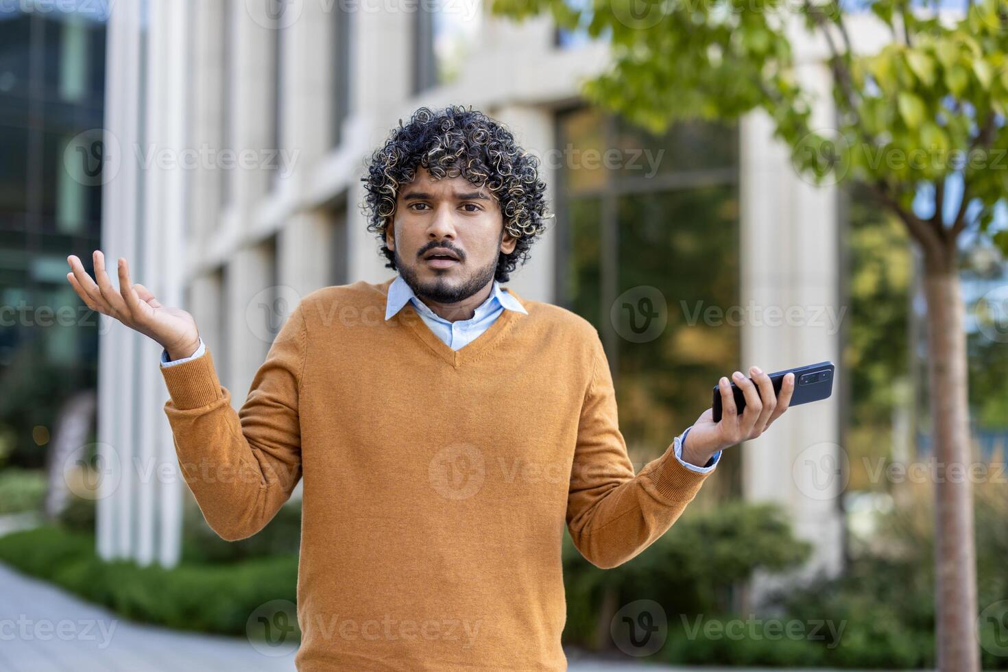 Young man with curly hair shrugs while holding smartphone, expressing confusion or uncertainty. photo