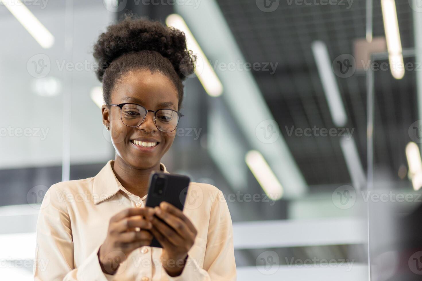 African American businesswoman smiling as she uses smartphone in modern office environment. Image captures professional interaction with digital technology, emphasizing cheerful mood photo