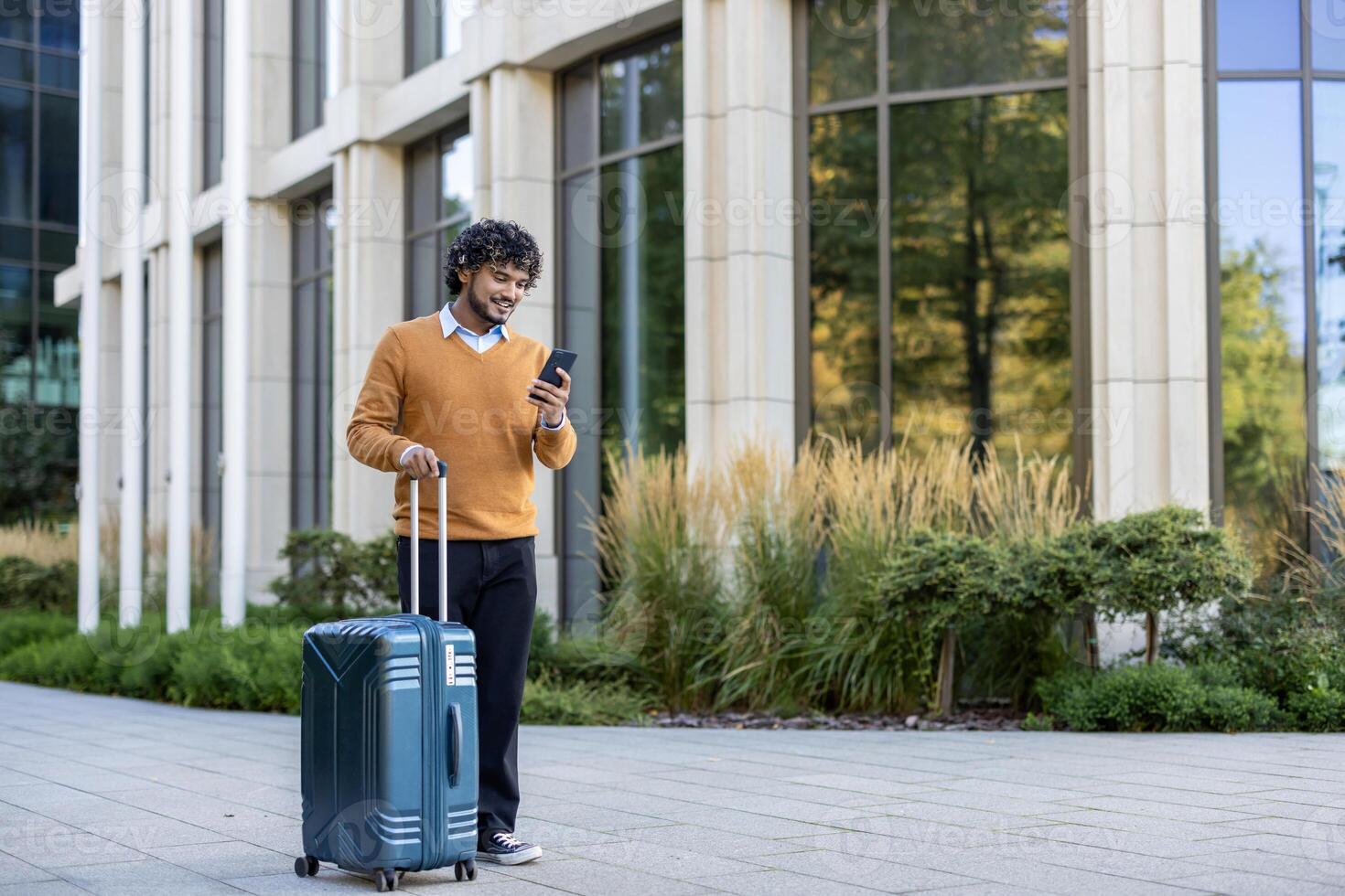 Smiling business traveler using smartphone while pulling suitcase in urban setting. Person wearing orange sweater walking outside office building. Captures concept of business travel photo