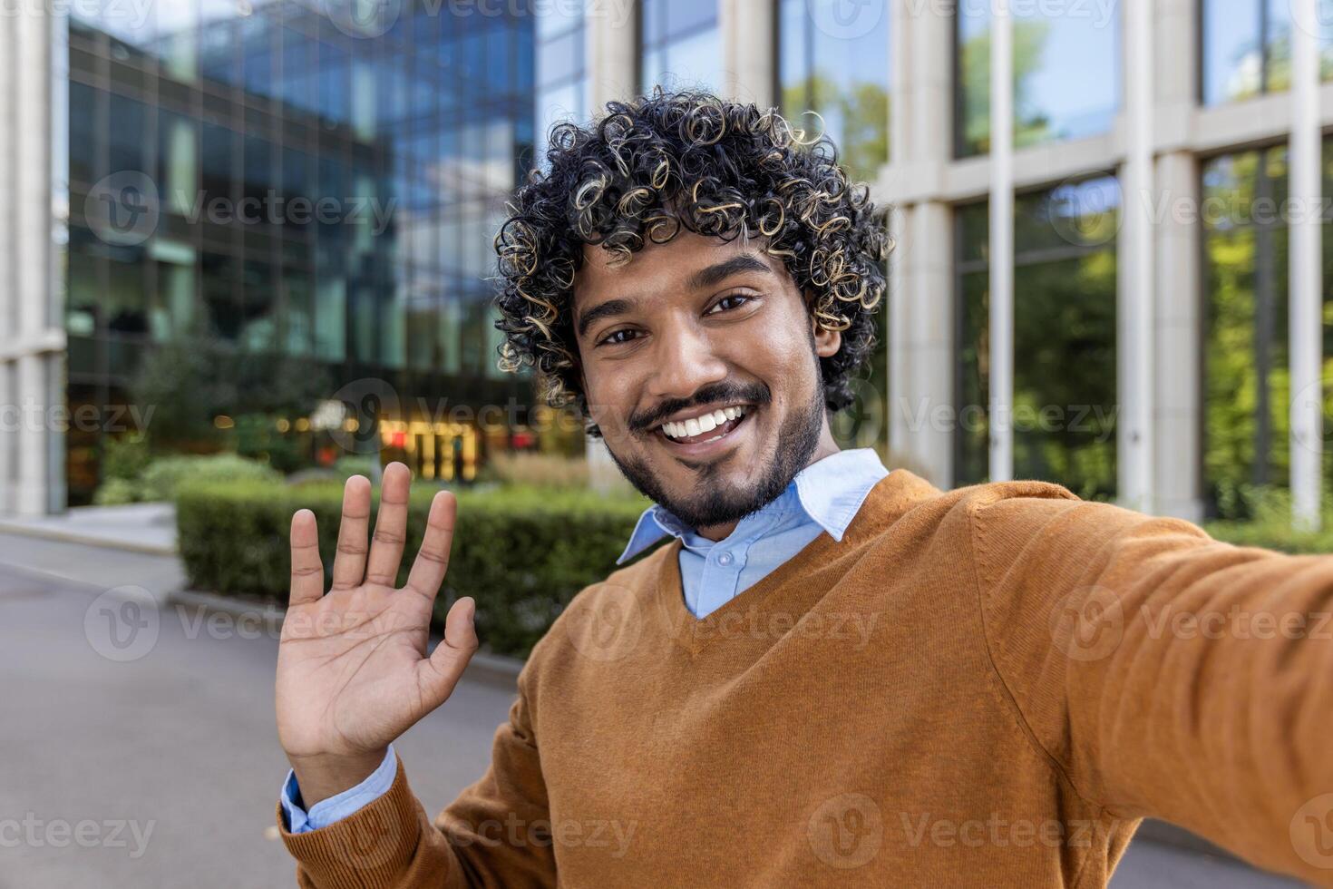 Cheerful man taking selfie outside modern office building. Man is smiling, waving, creating welcoming atmosphere. Bright sunny day enhances positive and professional mood. photo