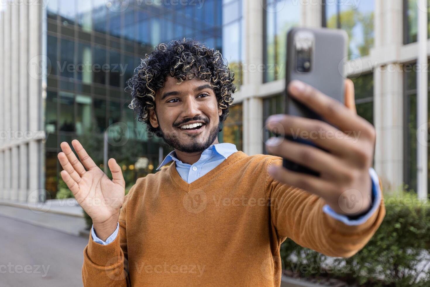 Cheerful man taking selfie with phone in city environment, wearing casual clothes. Captures joyful expression, waving to camera, experiencing happiness and connection. Ideal for themes of technology, communication, and modern lifestyle. photo