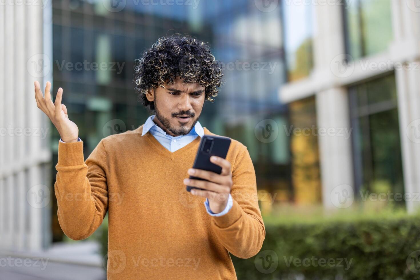Man with curly hair standing outside modern office building looks at phone with frustration. Wearing a brown sweater, he expresses confusion or dissatisfaction with technology issues. photo