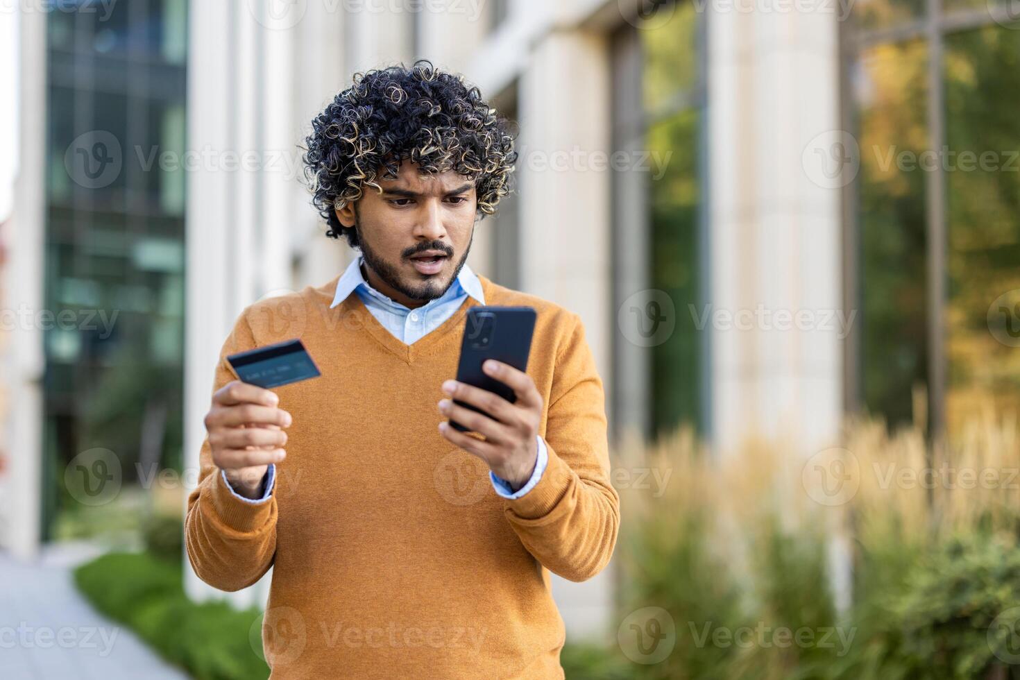 Concerned man using phone outdoors, holding credit card, experiencing trouble online banking or payment. Wearing casual attire, standing by modern building, showing expression of worry or confusion. photo