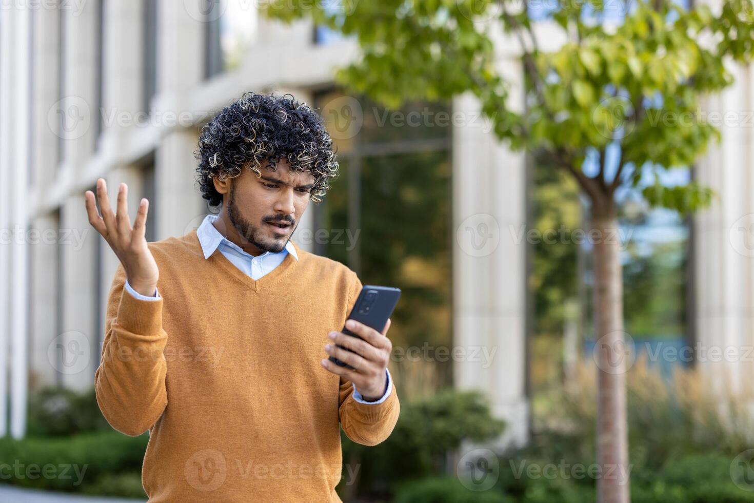 Young man feeling frustrated while looking at phone outside modern office building. He gestures with confusion, expressing annoyance. Wearing brown sweater, standing in urban environment under tree. photo