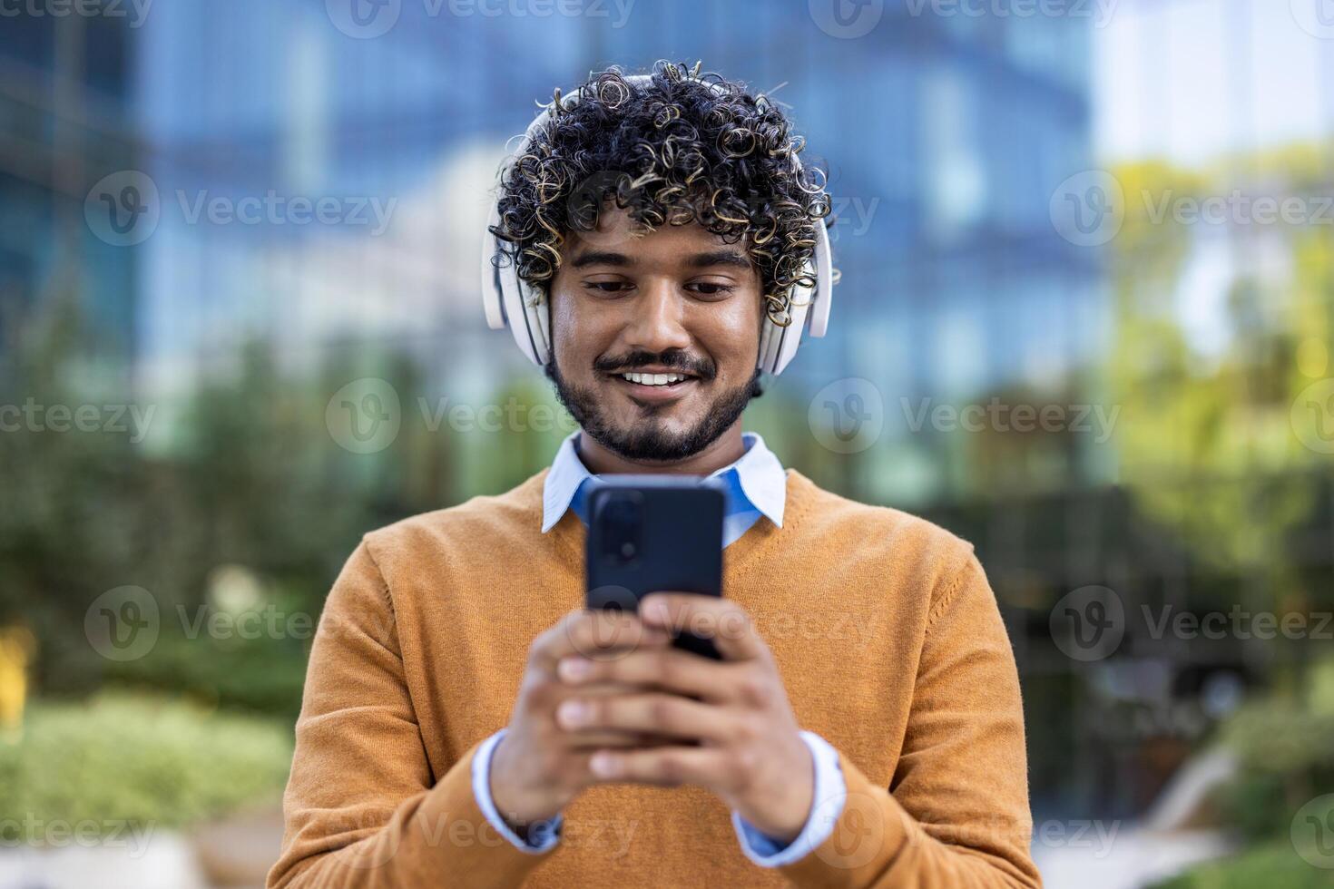 Portrait of smiling man wearing headphones, using smartphone outdoors in urban environment. Person enjoying music with phone. Capturing moment of happiness and connectivity, highlighting technology photo