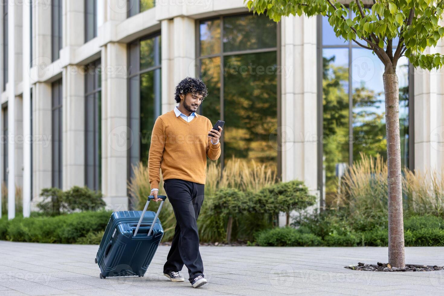 Man dressed in casual clothing walking with suitcase, using smartphone outside modern office building. Represents travel, technology, and connectivity in urban environment. photo