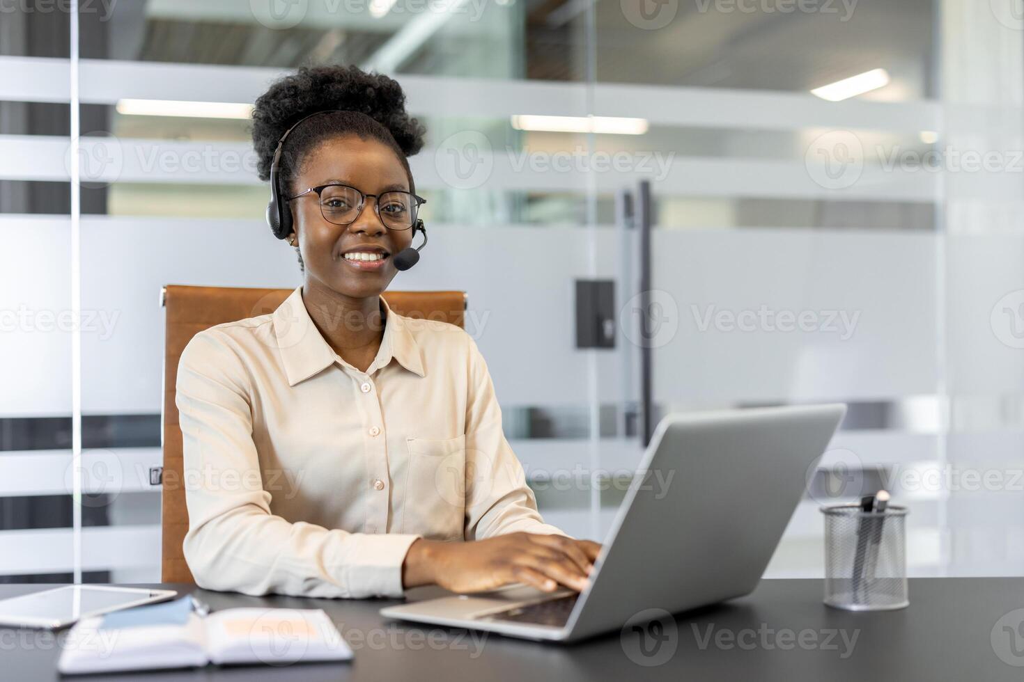 Confident African American businesswoman using laptop and headset for customer support. She is smiling, focused, and working efficiently in modern office setting. photo