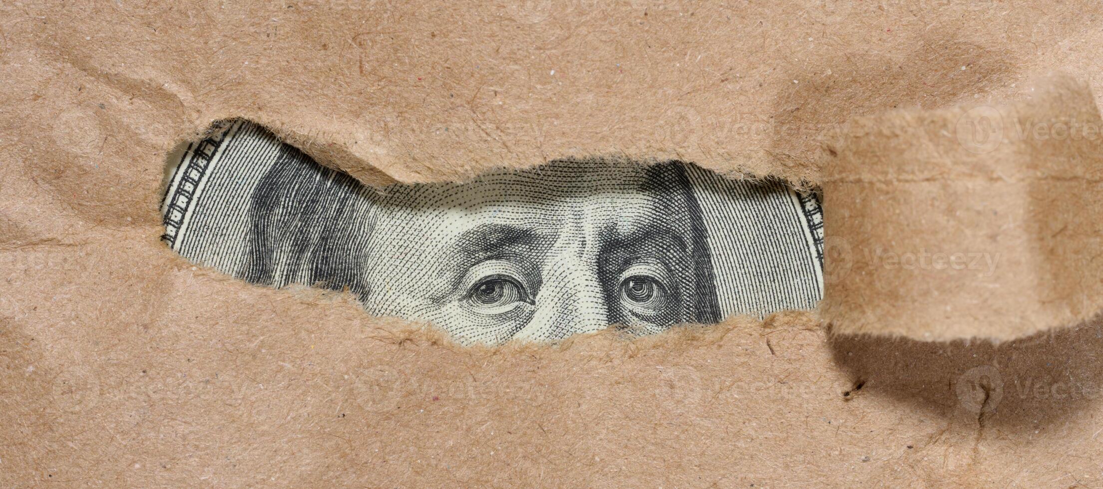 A piece of a hundred dollar bill is visible through a hole in the paper. Symbolism of hidden or concealed wealth, money that remains out of sight, or financial resources. photo