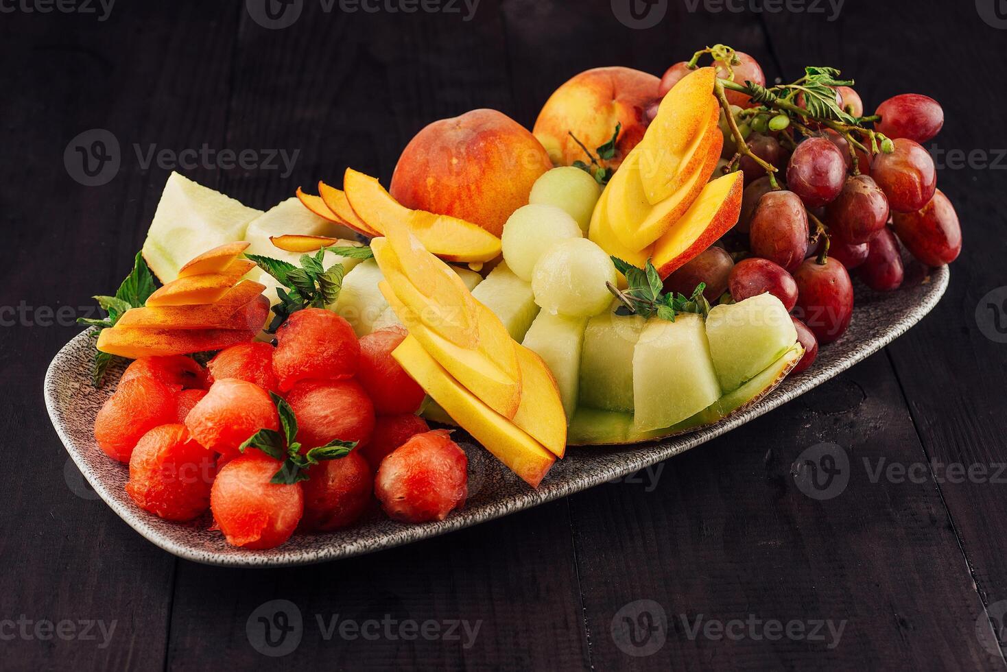 Oval plate overflowing with fresh watermelon, cantaloupe, peaches and grapes on dark wooden table photo