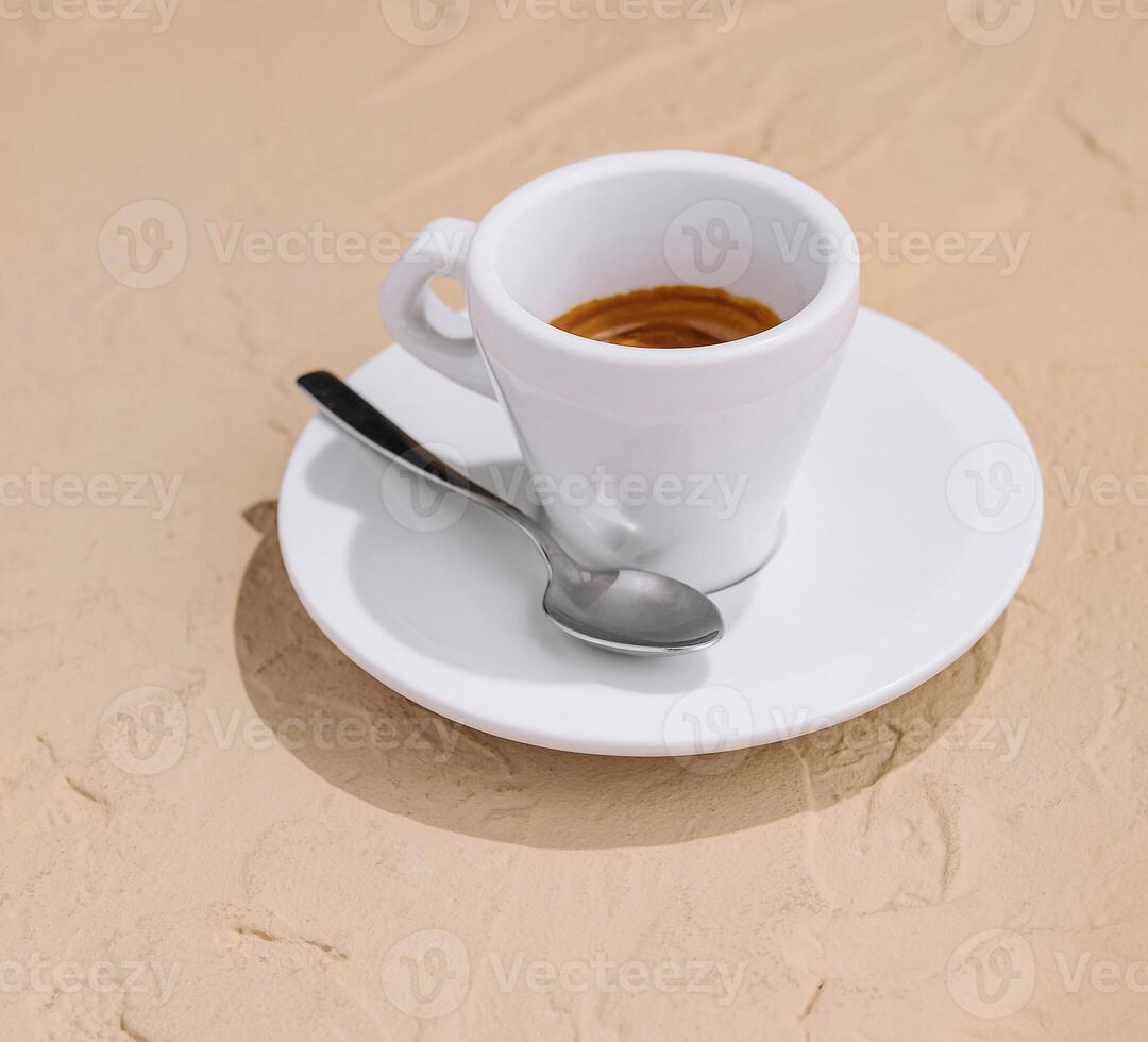 A white cup of espresso coffee on a sandy surface under soft sunlight photo