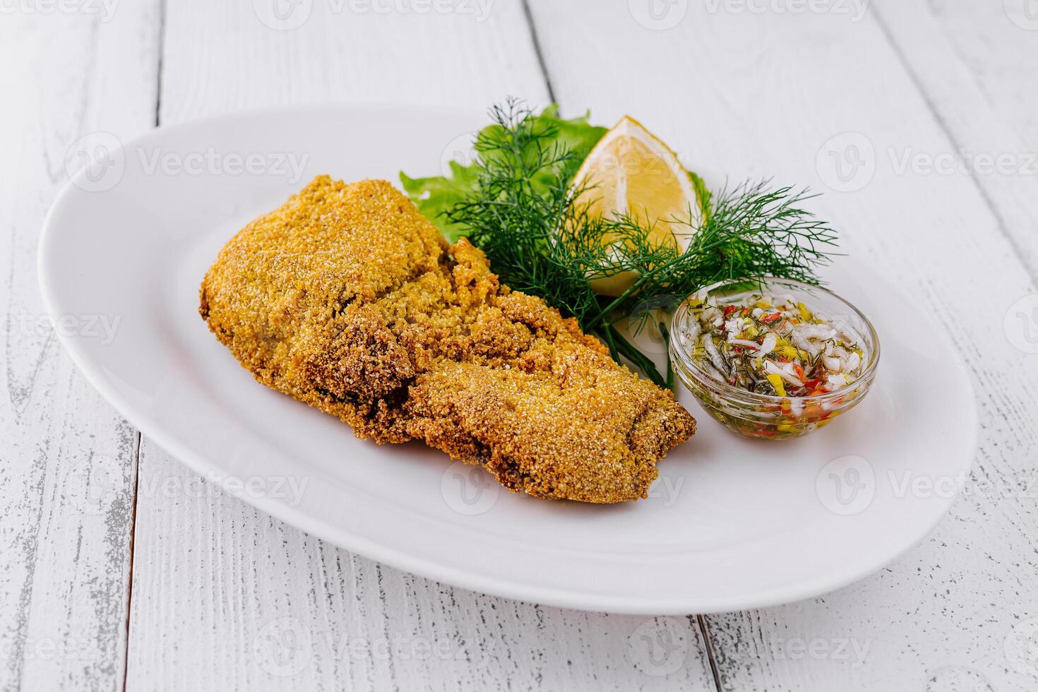 Breaded fish fillet being served on white plate with lemon and greens photo