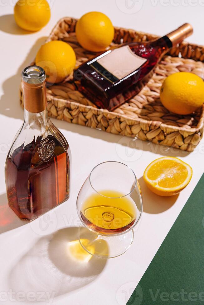 Bottle and glass of cognac with lemons on white and green background photo