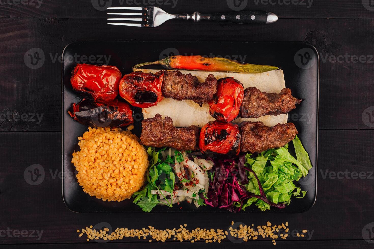 Turkish adana kebab with vegetables on black plate being served with bulgur photo
