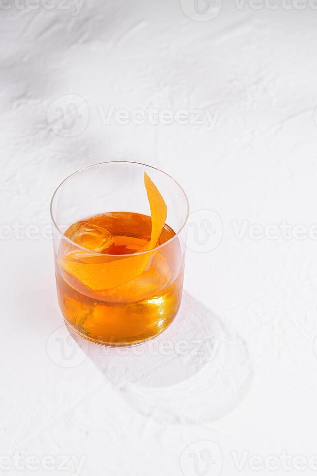 Glass with ice and orange peel is casting a shadow on white table photo