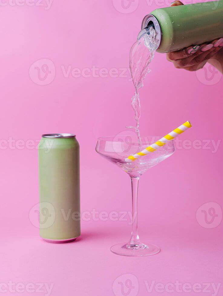Woman pouring drink from can into glass with straw on pink background photo