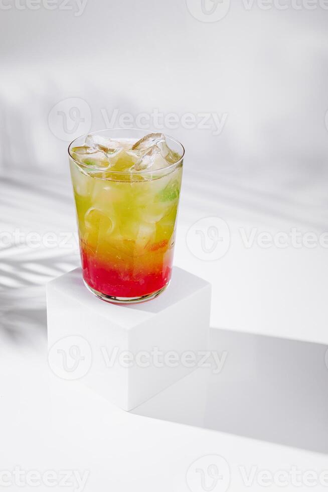Colorful layered cocktail standing on white cube with tropical shadow photo