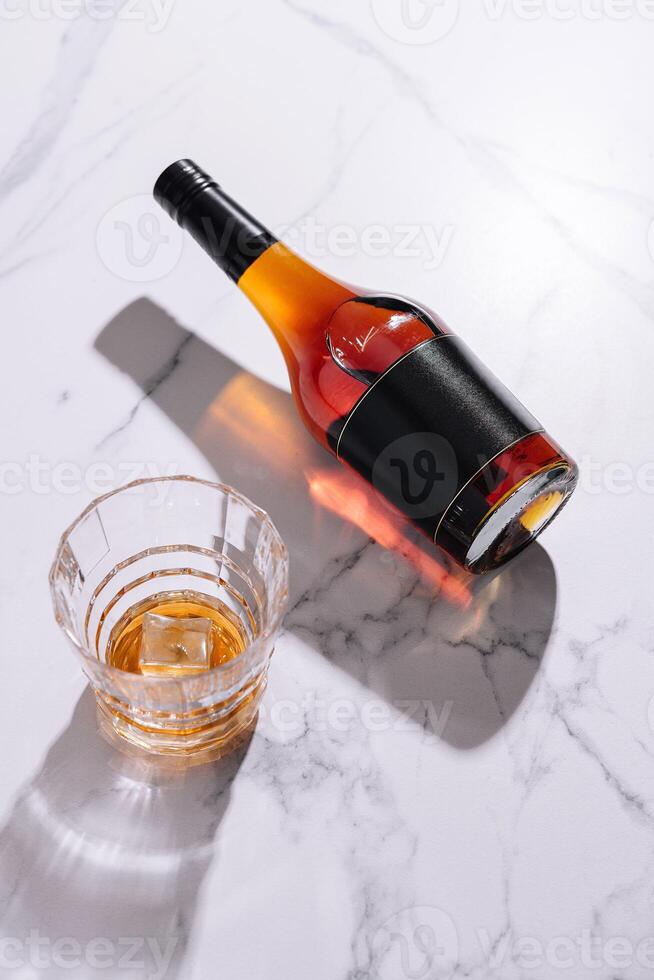 Bottle of liquor casting shadow near rocks glass with ice on marble photo