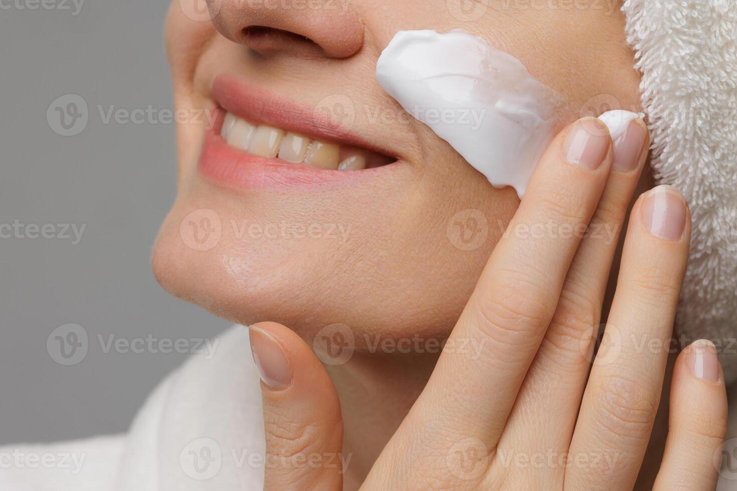 A woman applying cream to her face, smiling gently. She has a towel wrapped around her head and is in a relaxed setting, suggesting a skincare routine or spa treatment. photo