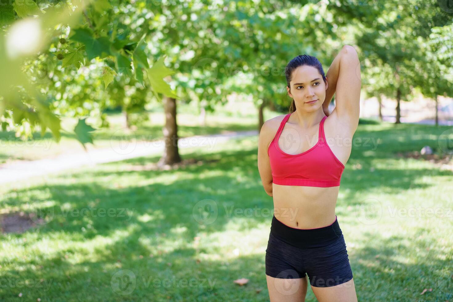 A woman in red sports bra and black shorts exercises outdoors, enjoying the summer sun in a beautiful park photo