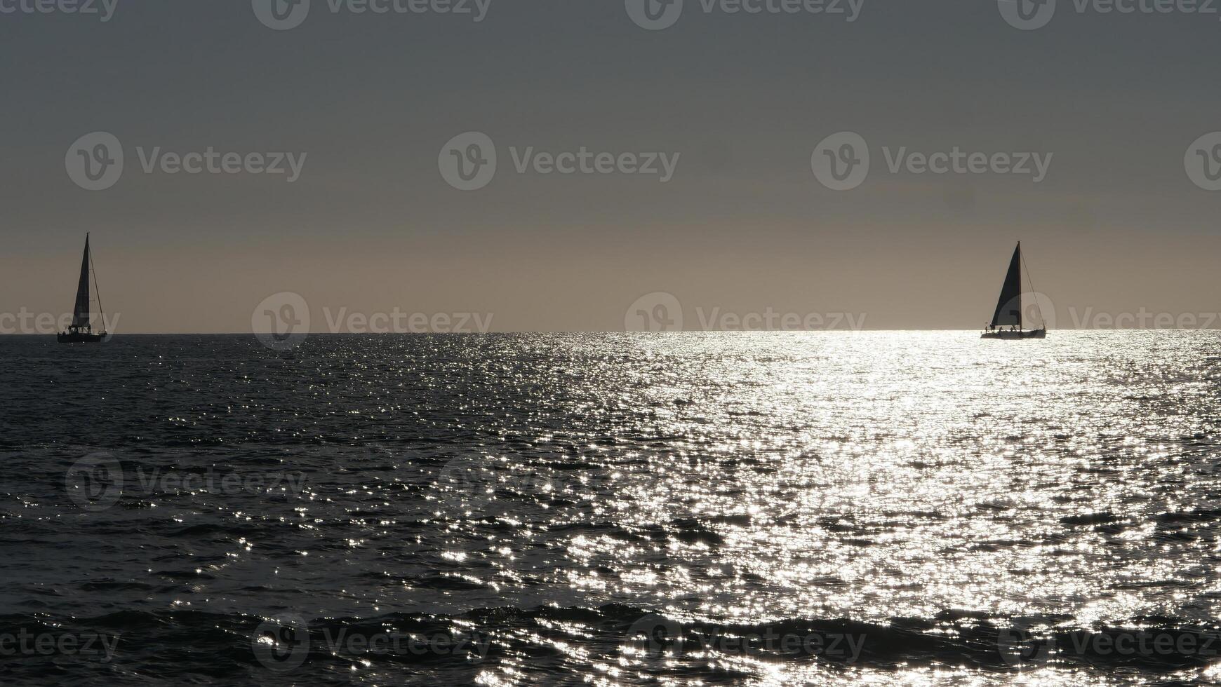 Two sailboats sailing away on calm ocean at sunset with sparkling water photo