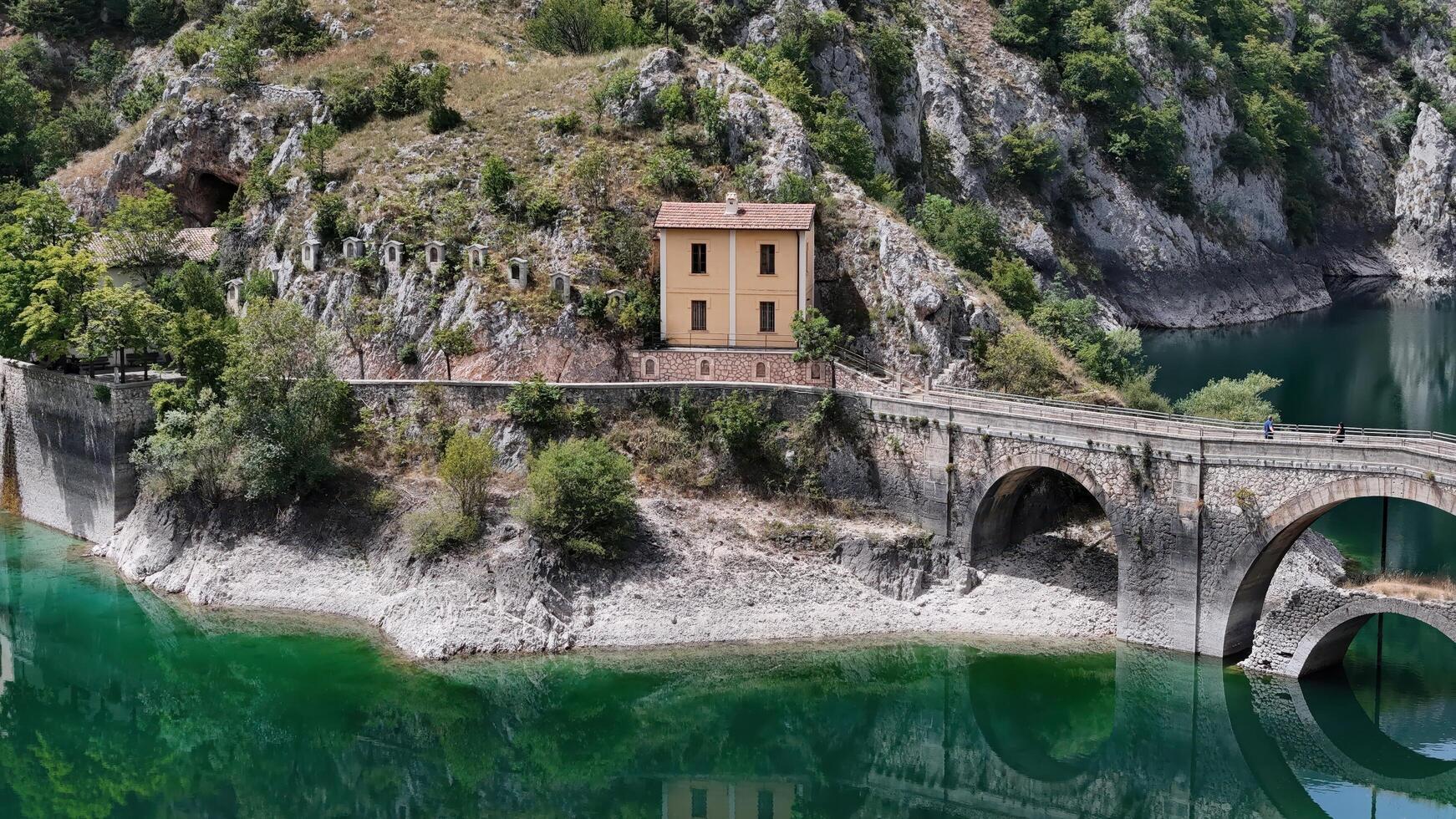 A quaint yellow house nestled between rocky hills and a serene emerald lake on a sunny afternoon photo