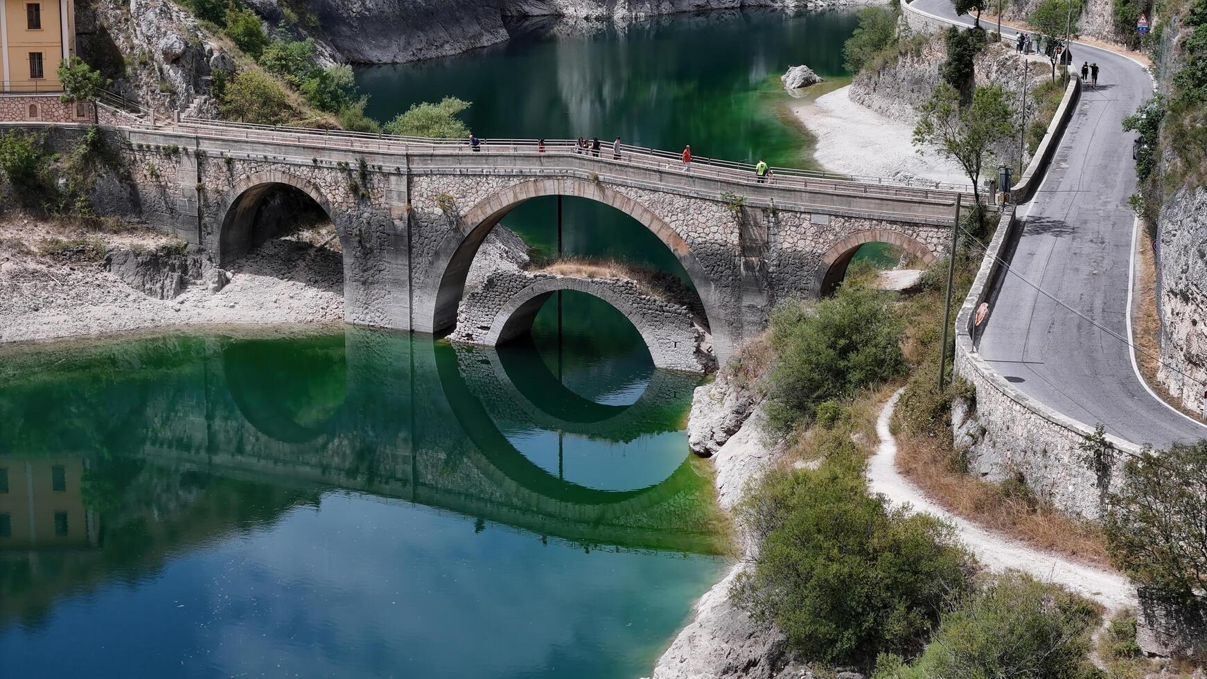 An ancient stone bridge arches gracefully over a vivid green river in Italy, surrounded by lush landscapes and a winding road photo