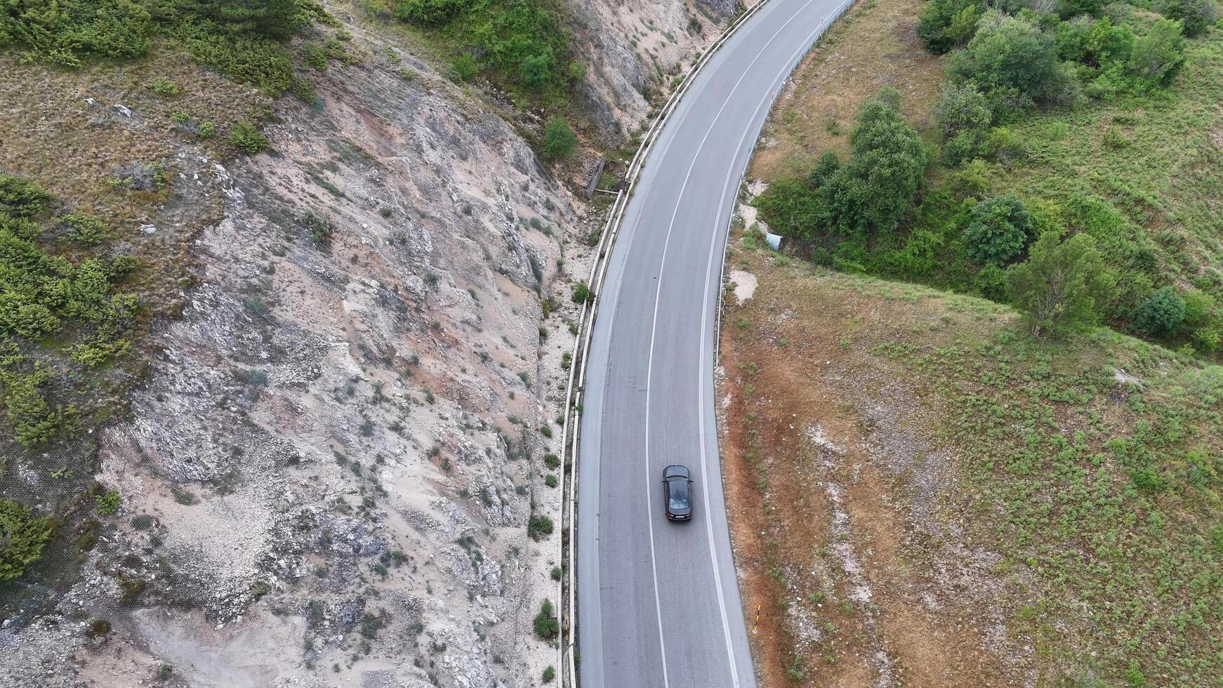 A solitary car navigates the winding road beside a rocky cliff in a scenic landscape on a calm, cloudy day photo