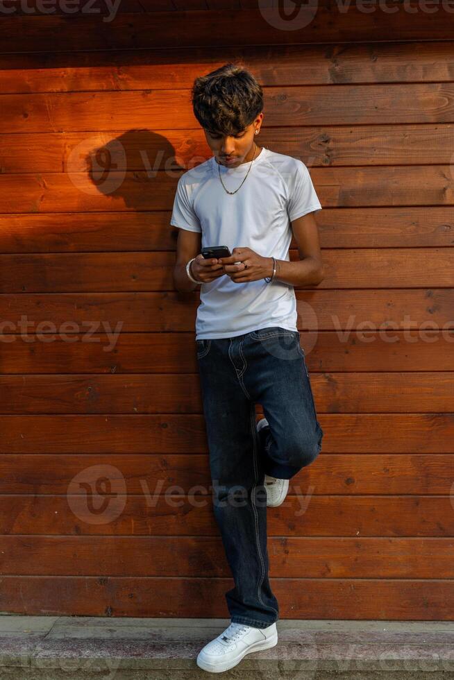 handsome teenage boy having fun using his cell phone leaning against a wooden plank wall photo