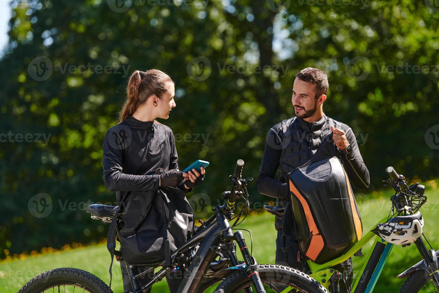 A sweet couple, equipped with bicycles and engrossed in coordinating their journey, checks their GPS mobile and watches while planning scenic routes in the park, seamlessly blending technology and photo