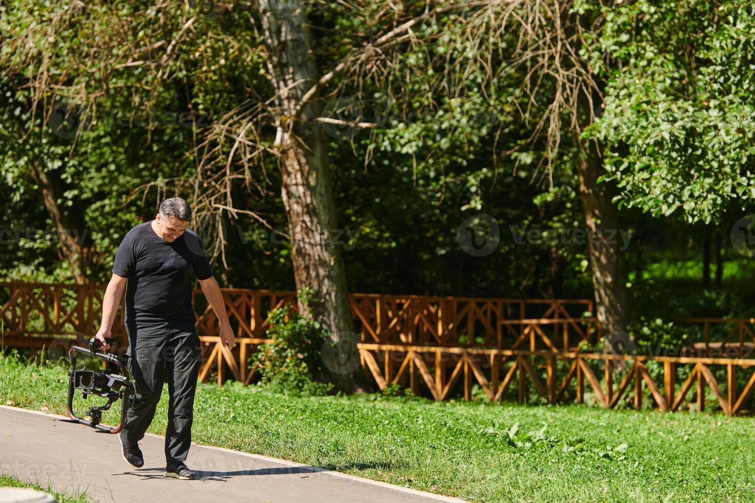 After completing a cinematic venture, a skilled videographer, adorned with camera equipment, leisurely strolls through the park, capturing the essence of post-production satisfaction and creative photo