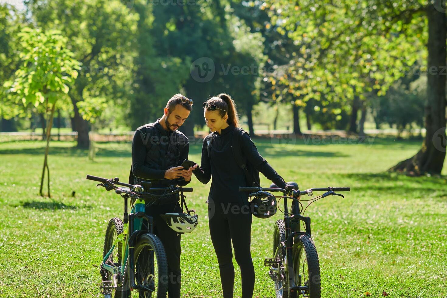 A sweet couple, equipped with bicycles and engrossed in coordinating their journey, checks their GPS mobile and watches while planning scenic routes in the park, seamlessly blending technology and photo