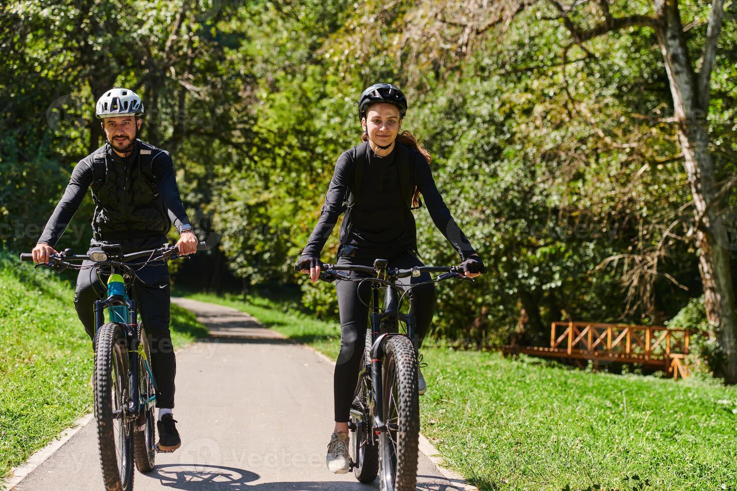 A blissful couple, adorned in professional cycling gear, enjoys a romantic bicycle ride through a park, surrounded by modern natural attractions, radiating love and happiness photo
